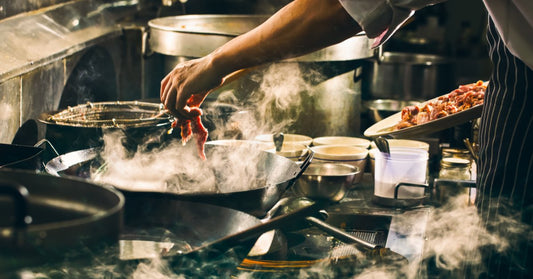 A close-up of a person in a white chef coat stirs food in a pan as steam rises from a stovetop in a dark kitchen.