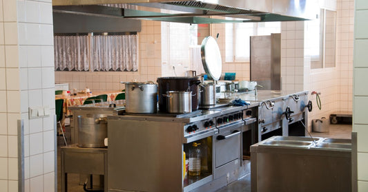 A large commercial kitchen with stainless steel counters, shelves, cookware, and bright overhead lights.